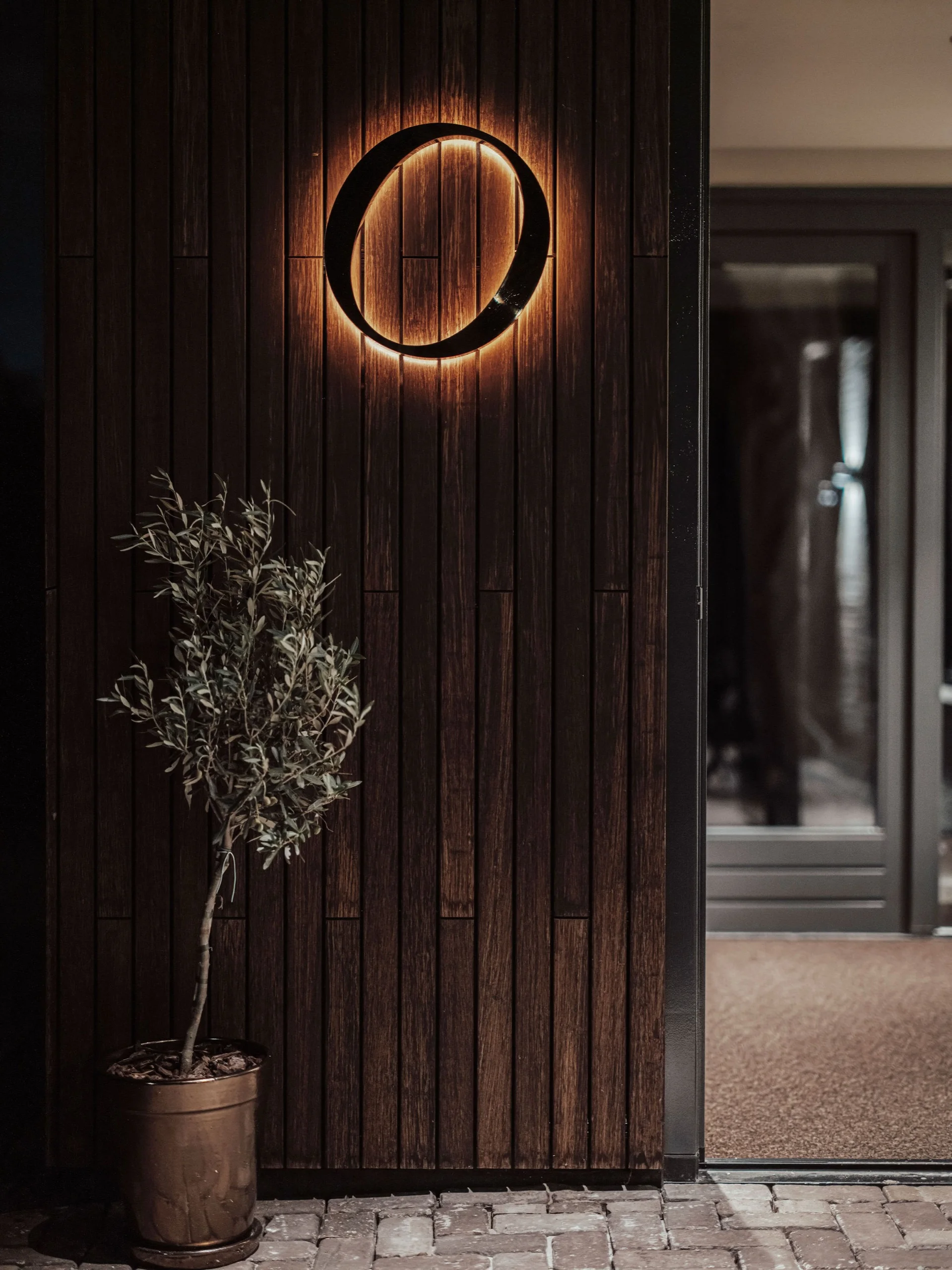 Verlicht rond logo op donkere houten gevel naast een olijfboompje bij de ingang van restaurant Olijf