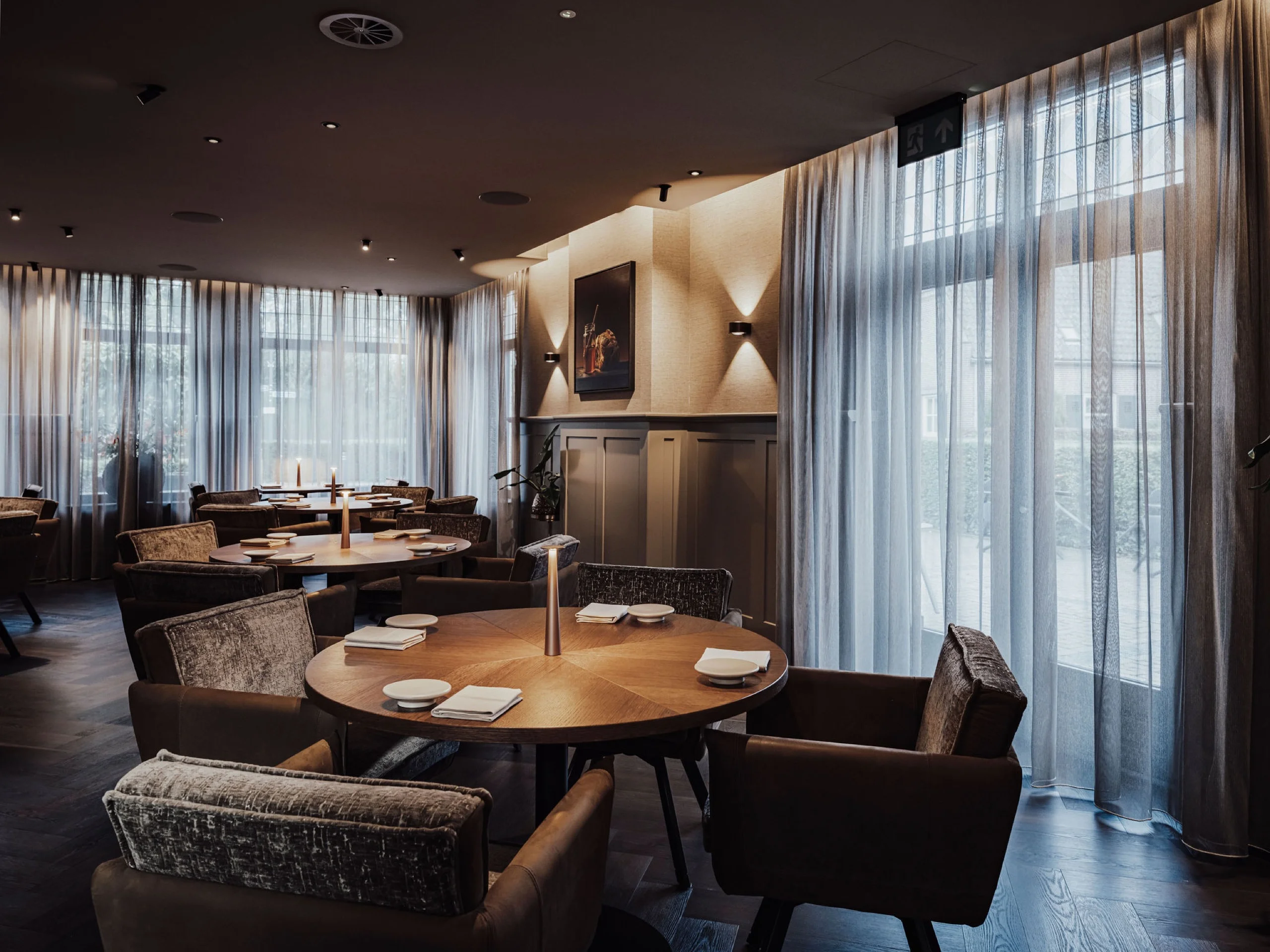 Sfeervol restaurantinterieur met ronde houten tafels, fluwelen fauteuils en grote ramen met vitrage